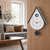 AMS wall clock on wooden surface, teardrop shape with light gray housing and black cuckoo