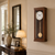 Wanduhr aus Holz mit rundem Zifferblatt und Pendel im schmalen Wandgehäuse